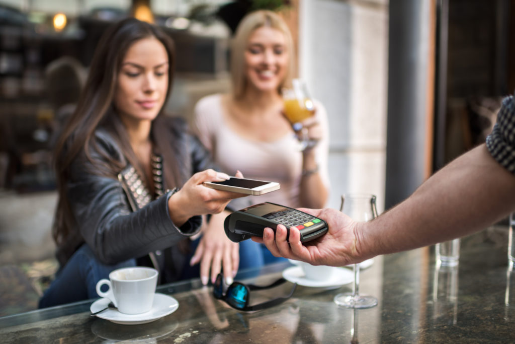 One of two female customers is holding mobile phone over credit card reader for contactless payment transaction in the coffee shop. Coffee cups and glasses are on the table.